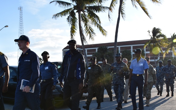A group of military and law enforcement personnel in various uniforms are walking together across a paved parking area or roadway. The scene appears to be a joint multinational exercise or official visit, with personnel wearing different service uniforms including what appears to be U.S. Coast Guard and other national military uniforms. The setting is a tropical coastal location with palm trees, a multi-story building, and a communications tower visible in the background.