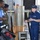 A U.S. Coast Guard officer in navy blue uniform is speaking with two foreign military personnel on the deck of a vessel. The individuals appear to be engaged in a briefing or exchange, likely as part of an international military cooperation or training event. One visitor wears a light blue uniform with a 'Barbados' patch, and the other wears camouflage military attire.