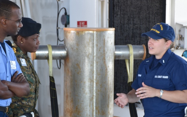 A U.S. Coast Guard officer in navy blue uniform is speaking with two foreign military personnel on the deck of a vessel. The individuals appear to be engaged in a briefing or exchange, likely as part of an international military cooperation or training event. One visitor wears a light blue uniform with a 'Barbados' patch, and the other wears camouflage military attire.
