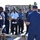A military or coast guard instructor is conducting an outdoor training briefing for a group of uniformed personnel from multiple nations or services. The instructor, facing away from the camera, appears to be demonstrating or explaining equipment — specifically a pipe or valve assembly mounted on a stand in the foreground. The setting appears to be a waterfront or port facility with tropical vegetation and institutional buildings visible in the background.