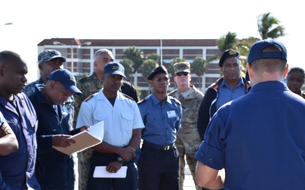A military or coast guard instructor is conducting an outdoor training briefing for a group of uniformed personnel from multiple nations or services. The instructor, facing away from the camera, appears to be demonstrating or explaining equipment — specifically a pipe or valve assembly mounted on a stand in the foreground. The setting appears to be a waterfront or port facility with tropical vegetation and institutional buildings visible in the background.
