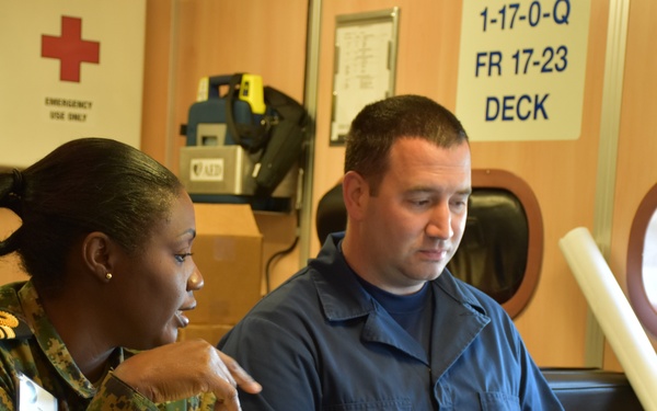 Two military personnel are seated at a table aboard what appears to be a naval vessel, collaborating over a ruggedized laptop computer. A woman in camouflage uniform with gold rank insignia is pointing at the screen while a man in blue coveralls operates the keyboard. The setting includes visible safety equipment and naval vessel markings on the wall.