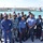 A group of approximately 16-18 military and law enforcement personnel in various uniforms are posed for a group photo on a dock or pier. In the background, two U.S. Coast Guard vessels are visible moored at what appears to be a Coast Guard station or naval facility. The gathering appears to be a multinational or joint-service event, with personnel wearing different national and branch uniforms.