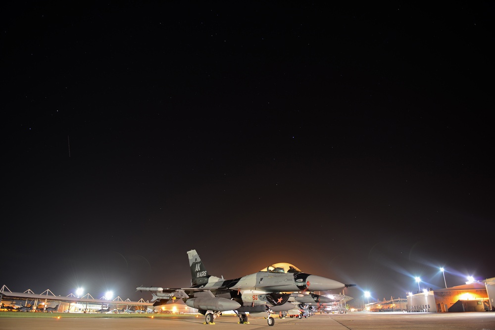 A military fighter jet, identified as an F-16 Fighting Falcon, is parked on a flight line at night under a clear, star-filled sky. The aircraft bears 'AK' tail markings indicating it belongs to an Alaska-based unit, with '18 AGRS' visible on the tail, identifying it as part of the 18th Aggressor Squadron. The scene is illuminated by ground-level floodlights from surrounding base infrastructure.