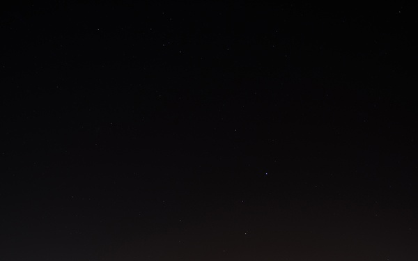 A military fighter jet, identified as an F-16 Fighting Falcon, is parked on a flight line at night under a clear, star-filled sky. The aircraft bears 'AK' tail markings indicating it belongs to an Alaska-based unit, with '18 AGRS' visible on the tail, identifying it as part of the 18th Aggressor Squadron. The scene is illuminated by ground-level floodlights from surrounding base infrastructure.