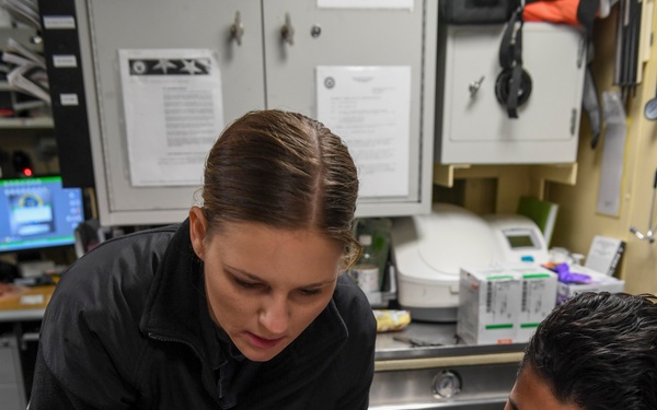 A woman in a black jacket is applying simulated wound makeup to a man's forearm using a small brush. The setting appears to be a medical or training facility, with moulage (casualty simulation) supplies visible on the table including a palette of theatrical makeup colors, powders, and applicators. This appears to be a medical training exercise where realistic wound simulation is being created for practice purposes.