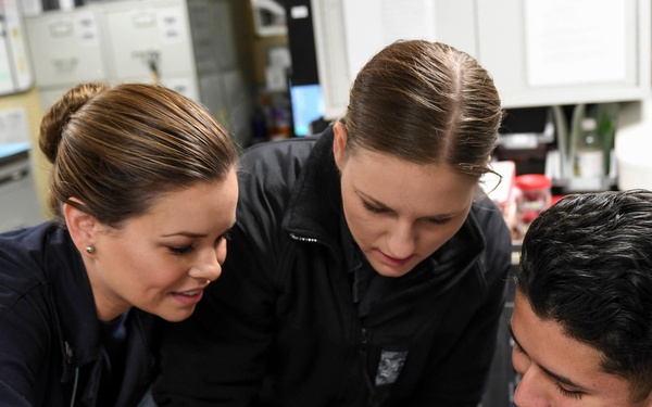 Two women in dark uniforms are attending to a wound or injury on a young man's arm in what appears to be a medical or training facility. One woman is applying direct pressure or treatment to a bloody wound on the man's forearm while the other observes closely. The setting includes medical supplies and storage cabinets visible in the background.