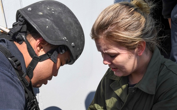 A military or naval training exercise depicting a simulated casualty scenario. A person wearing a tactical helmet and gloves is attending to a simulated wound on another person's leg. The 'wound' appears to be a realistic moulage (theatrical makeup/prosthetic) used for training purposes.