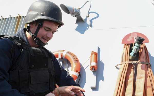 A military or law enforcement personnel wearing a tactical vest, ballistic helmet, and communications headset is performing what appears to be a medical aid or casualty care procedure on a person lying on a ship's deck. The individual's hands show visible red staining consistent with simulated or actual blood, suggesting a combat casualty care drill or real emergency response. The setting is clearly aboard a naval vessel, evidenced by life rings, fire hoses, and ship superstructure in the background.