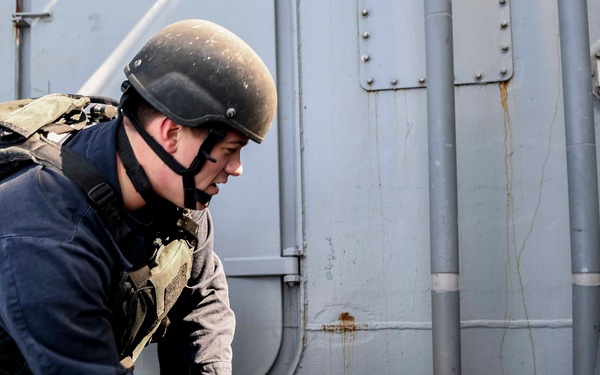 A military or law enforcement personnel wearing a helmet, tactical vest, and gloves is attending to a person lying on a surface, appearing to provide first aid or casualty care. The person on the ground is wearing olive/military-colored clothing and appears to have simulated injuries, suggesting this is likely a training exercise. The setting appears to be on or near a naval vessel based on the gray metal hull visible in the background.