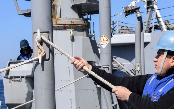 Naval personnel are working on the deck of a warship, handling thick rope lines during what appears to be a seamanship or line-handling evolution. The crew members are wearing blue safety vests and blue hard hats, indicating standard naval deck safety gear. The operation is taking place at sea under clear blue skies.