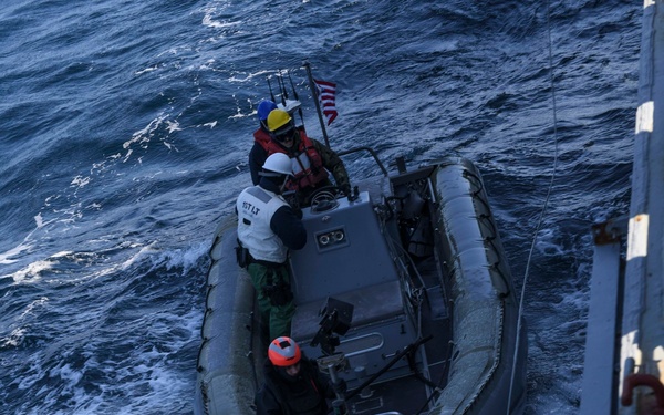 An aerial view of a rigid-hull inflatable boat (RHIB) operating in open ocean alongside a larger vessel. Three personnel wearing helmets and life vests are aboard the small craft, which displays a small American flag at the stern. The boat appears to be tethered or in close proximity to the parent vessel on the right side of the frame.
