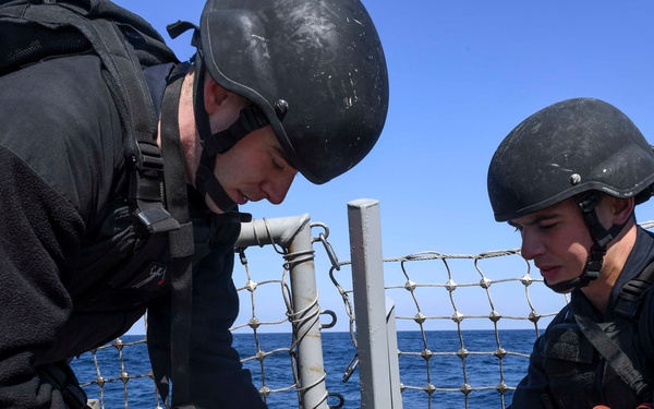 Two personnel in tactical gear are working on a rope or rigging system on the deck of a naval vessel at sea. Both individuals are wearing black helmets, tactical gloves, and dark uniforms with body armor or load-bearing equipment. They appear to be securing or preparing equipment near a safety netting railing with open ocean visible in the background.