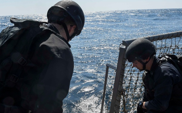 Two personnel dressed in black tactical gear and helmets are working on the deck of a naval vessel at sea. They appear to be handling equipment mounted on a red-framed structure near the ship's railing, with netting visible in the background. The ocean is clearly visible with sunlight reflecting off the water surface.