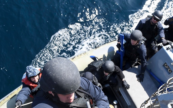 Military or law enforcement personnel are conducting a maritime boarding operation, climbing from a rigid inflatable boat (RIB) onto a larger vessel using ropes. The lead climber is wearing a ballistic helmet, tactical vest, and gloves marked 'FG', and is ascending using rope rigging. Several additional personnel in similar tactical gear remain in the RIB below, suggesting a visit, board, search, and seizure (VBSS) exercise or operation.
