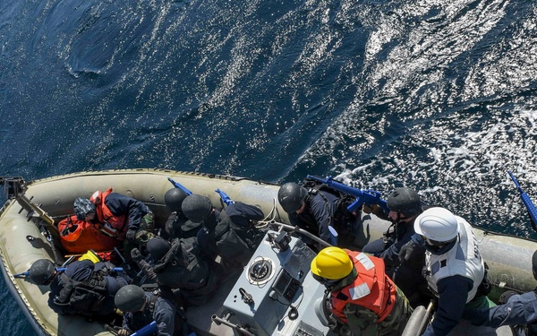 An aerial view of a rigid-hull inflatable boat (RHIB) carrying approximately 10-12 personnel on open ocean water. Several individuals are wearing tactical gear and helmets with blue-marked training weapons, while others wear orange life vests and colored hard hats, suggesting a maritime boarding or visit, board, search, and seizure (VBSS) training exercise. An American flag is visible at the stern of the vessel.