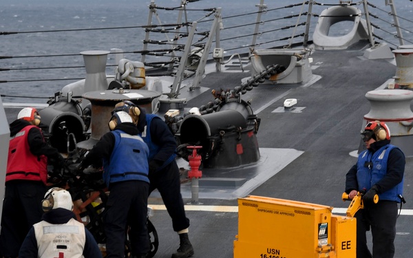 Naval personnel are conducting operations on the deck of a U.S. Navy warship at sea. Crew members in blue vests and hearing protection are working around deck equipment, while a yellow electric pallet jack marked 'USN 16-10646' sits on the flight/working deck. A crew member wearing a white vest with a Red Cross emblem is also present, indicating a medical or safety observer role.