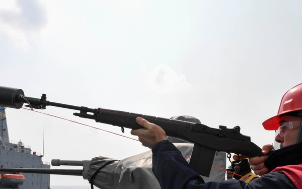 A naval sailor wearing a red hard hat (numbered 56) and red safety vest is firing a line-throwing rifle to establish a connection between two ships during an underway replenishment (UNREP) operation. A red messenger line is visibly attached to the rifle and being projected toward a large gray auxiliary/replenishment vessel positioned alongside. This is a standard military at-sea replenishment procedure used to transfer fuel, supplies, or personnel between ships.