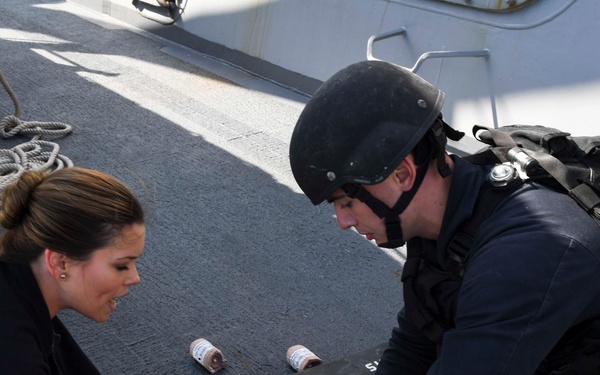 The image depicts what appears to be a military or naval medical training exercise on the deck of a ship. Two personnel in dark uniforms are attending to a third person lying on the deck, simulating a casualty scenario. One individual wears tactical gear including a helmet, while another has simulated blood on their hands, consistent with combat casualty care training.