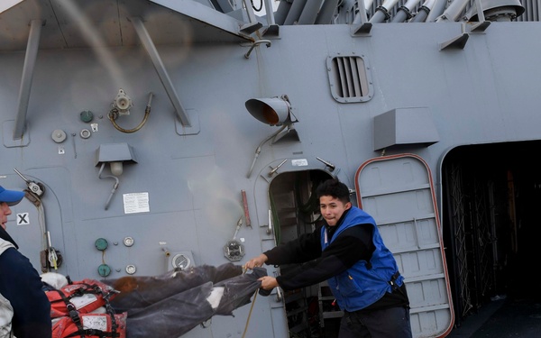 Two naval personnel are working on the deck of a warship, pulling on what appears to be a large dark tarp or cover together. One sailor on the left wears a white vest marked 'R108' and a blue cap, while the other wears a blue jacket and is pulling a rope attached to the material. The scene takes place on the exterior deck of a gray military vessel with various shipboard equipment visible.