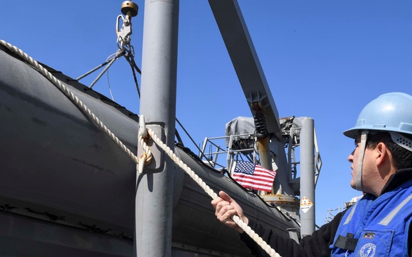 A sailor or naval crew member wearing a blue hard hat and blue flotation vest is handling a rope/line on the deck of a U.S. Navy vessel. A rigid inflatable boat (RIB) is suspended on a davit system above the deck. An American flag is visible in the background, confirming the vessel's U.S. Navy affiliation.