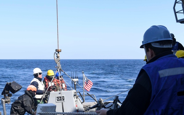 Naval personnel aboard a U.S. military vessel are conducting a small boat recovery or deployment operation. Crew members wearing hard hats and safety vests are managing lines and rigging to hoist or lower a rigid inflatable boat (RIB) using a crane/davit system. An American flag is visible on the small boat, indicating U.S. Navy affiliation.