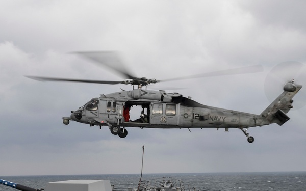 A U.S. Navy MH-60 Seahawk helicopter (marked '12' and 'NAVY') hovers over the flight deck of a U.S. Navy destroyer, appearing to conduct a vertical replenishment or personnel transfer operation. Deck crew members in colored safety vests are visible on the ship's deck, with a 5-inch deck gun prominently displayed in the foreground adorned with an American flag. The scene takes place at sea under overcast skies with moderate wave conditions.