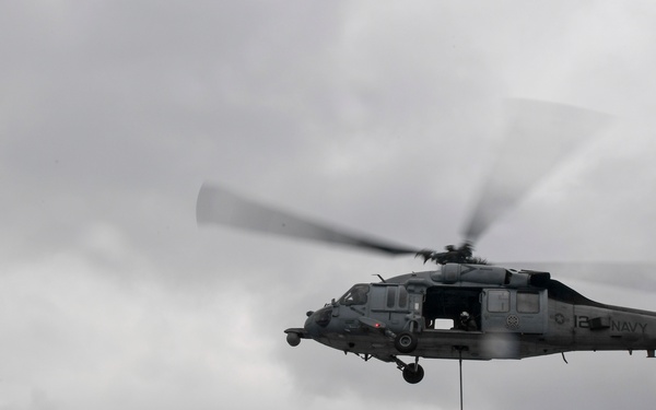 A U.S. Navy helicopter, marked 'NAVY' with tail number 12, hovers over the deck of a naval warship conducting a vertical replenishment (VERTREP) operation. A cargo net loaded with supplies is suspended beneath the helicopter via a hoist line. Personnel in blue vests are visible on the ship's deck near a naval gun mount decorated with an American flag.