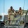 Two U.S. Air Force personnel in MultiCam/OCP camouflage uniforms are seated atop a HMMWV (Humvee) military vehicle, appearing relaxed and engaged in casual conversation. The vehicle is marked 'US AIR FORCE 05L 00420' and displays green camouflage paint. Concertina wire is visible in the foreground, suggesting a secured perimeter or staging area.