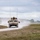 A convoy of military HMMWV (Humvee) vehicles is traveling along a dusty gravel road through an open, flat training area. The lead vehicle is prominently visible in the foreground with at least two additional vehicles following behind, all kicking up dust clouds. The setting appears to be a military training installation with open grassland and tree lines in the background.