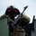A military soldier is positioned in the turret or hatch of a military vehicle, using large binoculars with red-tinted lenses to observe a distant area. The soldier is wearing full combat gear including a camouflage uniform and helmet with mounted equipment. A mounted machine gun is visible in the foreground, suggesting a defensive or reconnaissance posture.