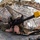 A soldier in camouflage uniform and helmet is positioned in a prone firing stance beneath a camouflage netting shelter, aiming a rifle equipped with a yellow MILES (Multiple Integrated Laser Engagement System) training device on the muzzle. The fisheye lens perspective exaggerates the barrel length and creates a wide-angle distortion of the scene. This appears to be a military training exercise based on the MILES gear and the controlled environment.