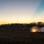 A military training exercise is captured at dusk showing a controlled detonation or explosive charge going off near a concertina wire perimeter. The scene depicts a forward operating base or field camp in the background with military tents and a communications antenna tower silhouetted against a sunset sky. The explosion produces visible sparks, flame, and smoke consistent with a breaching charge or simulated ordnance detonation.