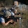 Military personnel are shown in a prone firing position on a grassy field, aiming rifles downrange. The foreground soldier wears ACU (Army Combat Uniform) digital camouflage helmet and tactical vest over a civilian-style shirt. Multiple soldiers are visible in a line extending into the background, suggesting a coordinated training exercise or range qualification event.