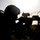 A silhouetted soldier in a prone firing position aims a rifle equipped with optical sights toward the right of the frame. The image is backlit by a bright sun, creating a strong silhouette effect against a pale sky. Coiled razor/concertina wire is visible in the background, indicating a defensive perimeter or training environment.