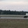 A military service member in camouflage uniform and full gear walks a dark-colored military working dog (MWD) on a long leash along what appears to be an airfield perimeter road. The scene is set at dusk or dawn, with a treeline in the background and a military building visible to the right. Concertina wire is visible in the foreground, indicating a secured military installation.