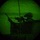 Image captured through a night vision device showing a circular green-tinted field of view. A silhouetted figure wearing a military helmet is visible aiming a rifle, positioned behind what appears to be a wall or barrier. The characteristic green phosphor glow and circular vignetting confirm this is night vision optic imagery.