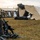 A military field camp scene showing a stack of rifles arranged in a traditional 'rifle stack' formation in the foreground, with soldiers in camouflage uniforms working on erecting or dismantling large military tents in the background. The setting appears to be an open grassy field at either dawn or dusk based on the diffuse lighting. This appears to be a training exercise or field operation setup.