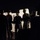 A nighttime scene showing a line of personnel holding large riot shields, backlit by a light source that illuminates the shields while leaving the figures largely in silhouette. The image appears to depict a law enforcement or security formation in a crowd control or riot control context. The high-contrast lighting creates a dramatic visual effect with dark surroundings.