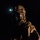 A person in full military or tactical gear is photographed against a completely dark background at night. They are wearing a gas mask, tactical helmet with a mounted flashlight illuminating their face, and camouflage uniform. The dramatic low-key lighting creates a stark, high-contrast image.