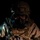 A soldier or military operator is photographed against a completely black background in dramatic low-key lighting. The individual is wearing full tactical gear including a gas mask, combat helmet, plate carrier vest, and camouflage uniform. The image appears to be a staged or studio-style military portrait emphasizing equipment and readiness.