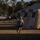 A person wearing a military-style helmet, tactical vest/backpack, and civilian athletic clothing (white t-shirt and blue shorts) is running across a dry grass field near large tan military-style tents. The setting appears to be a field training exercise or military base camp, with vehicles including a dark pickup truck visible in the background. The image is taken during golden hour lighting, creating strong shadows and warm tones.