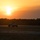 A military tactical vehicle, consistent with a HMMWV (Humvee), is positioned on what appears to be a flat open terrain or airfield tarmac during sunset or sunrise. The sun is low on the horizon, casting a warm orange and yellow glow across the sky and silhouetting a treeline in the background. The vehicle appears stationary or slowly moving along a paved surface.