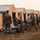 Military personnel in camouflage uniforms are setting up or breaking down a field camp consisting of multiple large tan and grey military tents arranged in a row. Soldiers are handling equipment including what appear to be folding cots or metal frame components stacked on the ground. The scene appears to take place during early morning or evening light in an open grassy field.