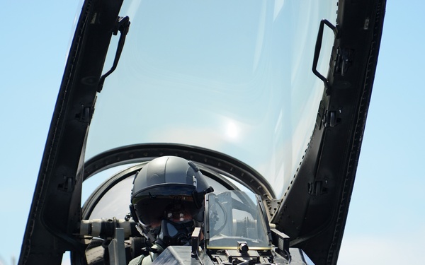 A military pilot is seated in the cockpit of a fighter jet, wearing a flight helmet and oxygen mask. The canopy is open, providing a close-up view of the pilot and the aircraft's instrument panel. A second aircraft is partially visible in the background on the left side.