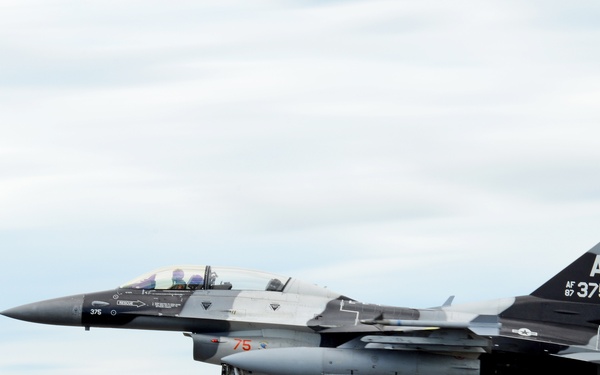 A U.S. Air Force F-16 Fighting Falcon with tail code 'AK 375' and serial number AF 87-375 is captured in low-altitude flight, likely on approach to landing with landing gear deployed. The aircraft bears Alaska Air National Guard markings, identifiable by the 'AK' tail code and the Alaska state silhouette emblem on the tail fin. The image uses a panning technique, creating motion blur in the background while keeping the aircraft relatively sharp.