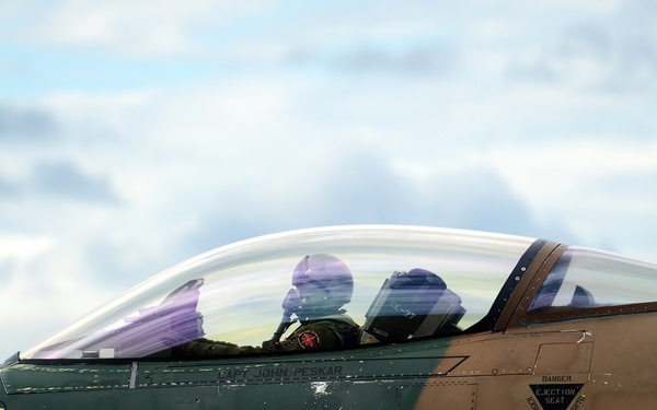 A two-seat fighter jet, likely an F-16D variant, is shown in close-up with two pilots seated in the cockpit under a clear canopy. The aircraft features a distinctive desert/jungle camouflage paint scheme in brown and green tones, with the number '286' on the nose and '86' on the fuselage. The name 'CAPT JOHN PESKAR' is stenciled below the cockpit, along with standard rescue and ejection seat markings.