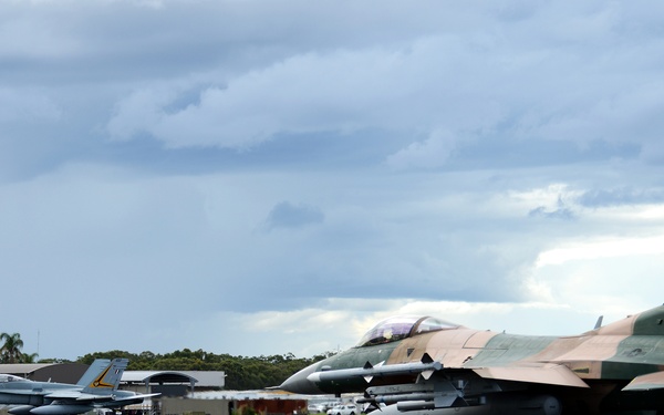 Two military jet aircraft are taxiing on a runway at a military airbase. The foreground aircraft is an F-16 Fighting Falcon in a distinctive camouflage paint scheme with tail code 'AK' and serial number '286', consistent with Alaska Air National Guard markings. A second grey jet aircraft, likely an F/A-18 Hornet, is visible in the background near a control tower with a yellow and black checkered pattern.