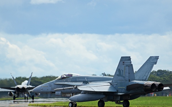 Two military jet aircraft are taxiing on a wet runway at what appears to be a military airbase or airshow. The foreground aircraft is a twin-engine fighter jet, likely an F/A-18 Hornet or Super Hornet, in a gray livery with visible markings. Lush green grass borders the taxiway, and a partly cloudy sky is visible in the background.