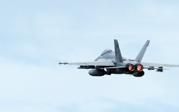 A twin-engine military jet fighter, consistent with an F/A-18 Hornet or Super Hornet, is captured in flight against a partly cloudy blue sky. The aircraft is viewed from a rear-quarter angle, with both engines visibly lit showing afterburner or exhaust glow. The image is sharp and well-exposed, clearly showing aircraft details including twin tail fins, wing-mounted stores, and cockpit canopy.